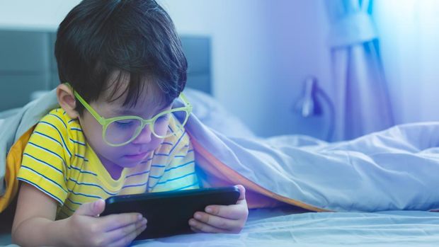 Ilustrasi anak main game detikhealth Cute little child watch movie on smartphone at bed. Dangers of blue light can damage eyes. Handsome little boy can be age related macular degeneration from blue light, wear eyeglasses since childhood