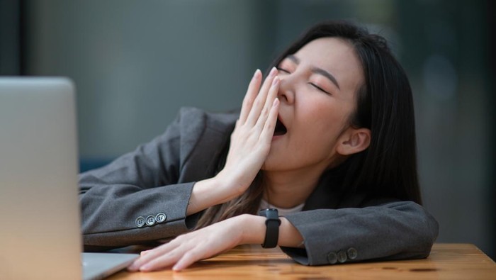 Young businesswoman yawning at a modern office desk in front of a laptop, covering her mouth out of courtesy