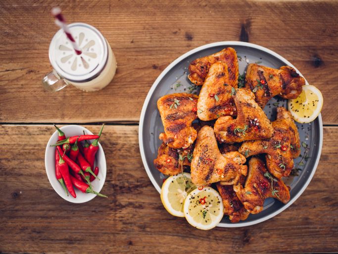 Overhead shot of a plate of spicy chicken wings with a bowl of fresh red chillies and a summer beverage on a vintage wooden surface