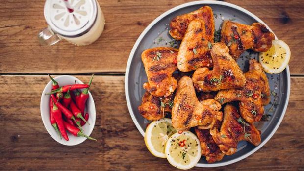 Overhead shot of a plate of spicy chicken wings with a bowl of fresh red chillies and a summer beverage on a vintage wooden surface