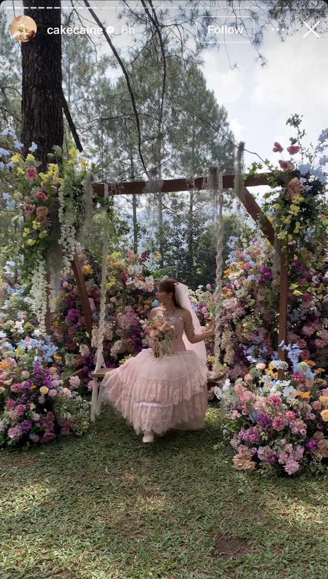 Roknya terdiri dari beberapa lapisan tulle dan renda yang bertumpuk, menciptakan volume dan nuansa klasik. Ia juga memakai veil putih panjang yang menjuntai lembut di belakang kepala. Foto: Instagram/@cakecaine
