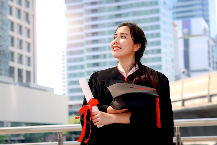 Happy smiling graduated student, young beautiful Asian woman holding square academic hat cap and certificate, standing with arms crossed at city with skyscraper building background, celebrating successful education on commencement graduation day when