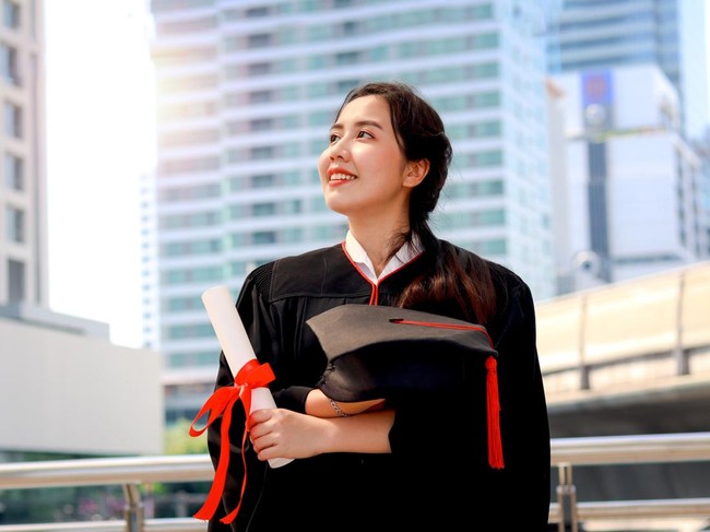Happy smiling graduated student, young beautiful Asian woman holding square academic hat cap and certificate, standing with arms crossed at city with skyscraper building background, celebrating successful education on commencement graduation day when finished studies for university or college.