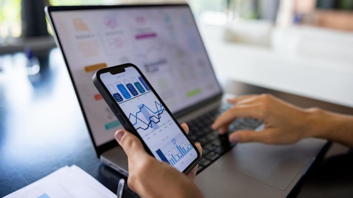 Close-up on a woman working at home and looking at the stock market behavior on her cell phone and the laptop - business concepts