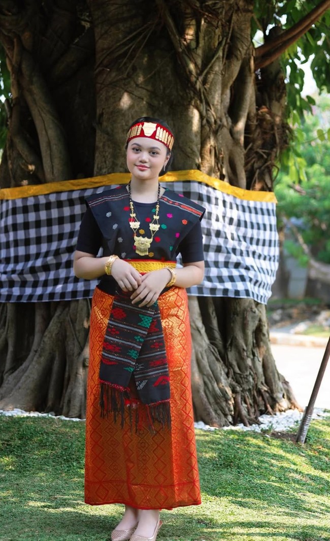 Amora Lemos tampil anggun mengenakan busana adat Batak lengkap, terdiri dari kain ulos berwarna hitam dengan motif merah, oranye, dan hijau, dipadukan dengan kain songket oranye keemasan yang dililit rapi di pinggang. Foto: Instagram/@amora_lemos05
