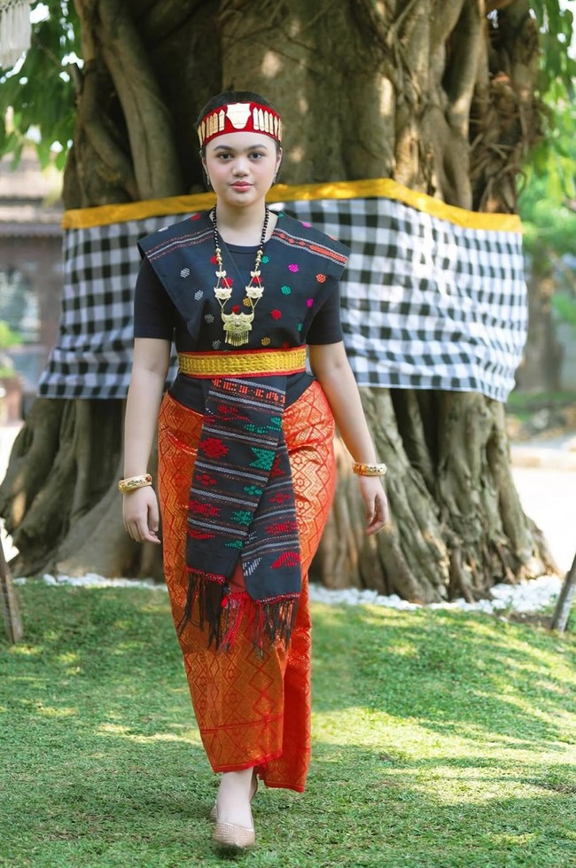 Penampilannya dipercantik dengan kalung emas besar berdesain khas, gelang emas di kedua tangan, serta manik-manik tradisional yang memperkuat nuansa adat. Foto: Instagram/@amora_lemos05