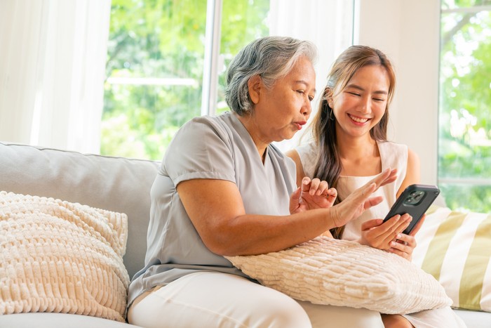 Happy Asian family senior mother and adult child spending time together at home. Daughter teach elderly mom using mobile phone application for online shopping and social media on sofa in living room.