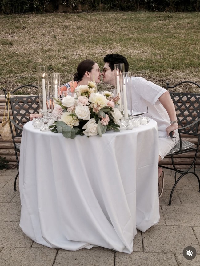 Meja bundar berbalut taplak putih ditata begitu elegan, dihiasi rangkaian bunga segar, lilin tinggi dalam tabung kaca, dan perlengkapan makan fine dining yang menciptakan suasana hangat dan romantis. Foto: Instagram/@lunamaya