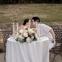 Meja bundar berbalut taplak putih ditata begitu elegan, dihiasi rangkaian bunga segar, lilin tinggi dalam tabung kaca, dan perlengkapan makan fine dining yang menciptakan suasana hangat dan romantis. Foto: Instagram/@lunamaya