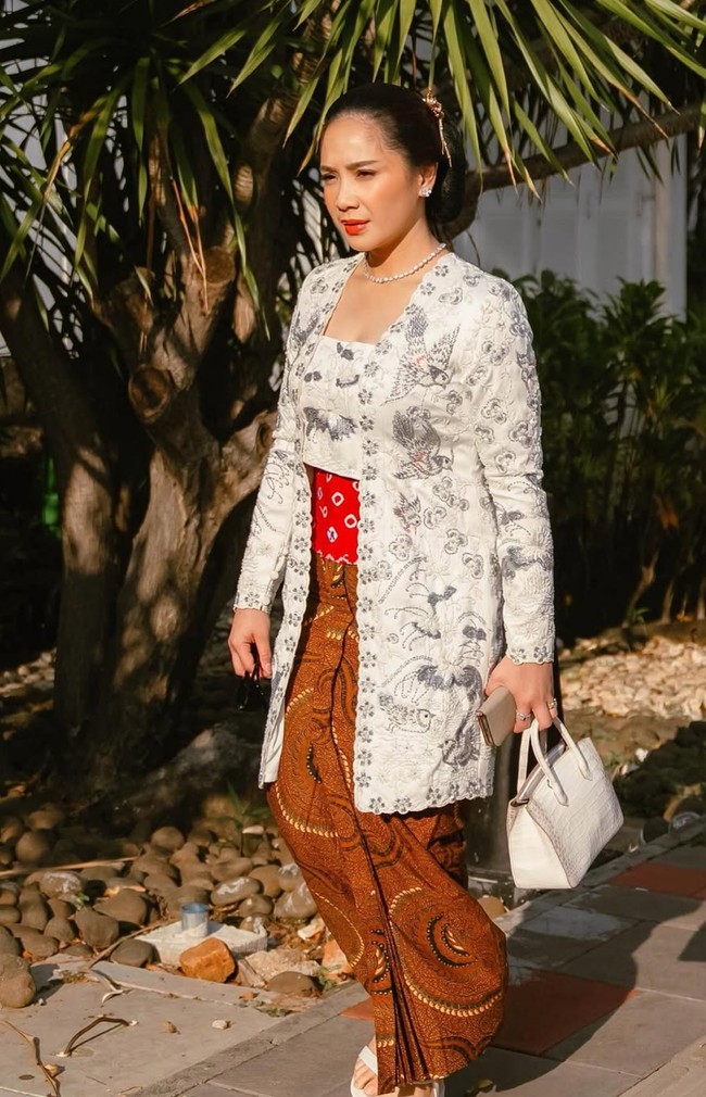 Nagita tampil anggun mengenakan kebaya brokat putih dengan detail bordir halus. Kebaya ini dipadukan dengan stagen merah bermotif, memberikan sentuhan kontras yang cantik. Foto: Instagram/@raffinagita1717