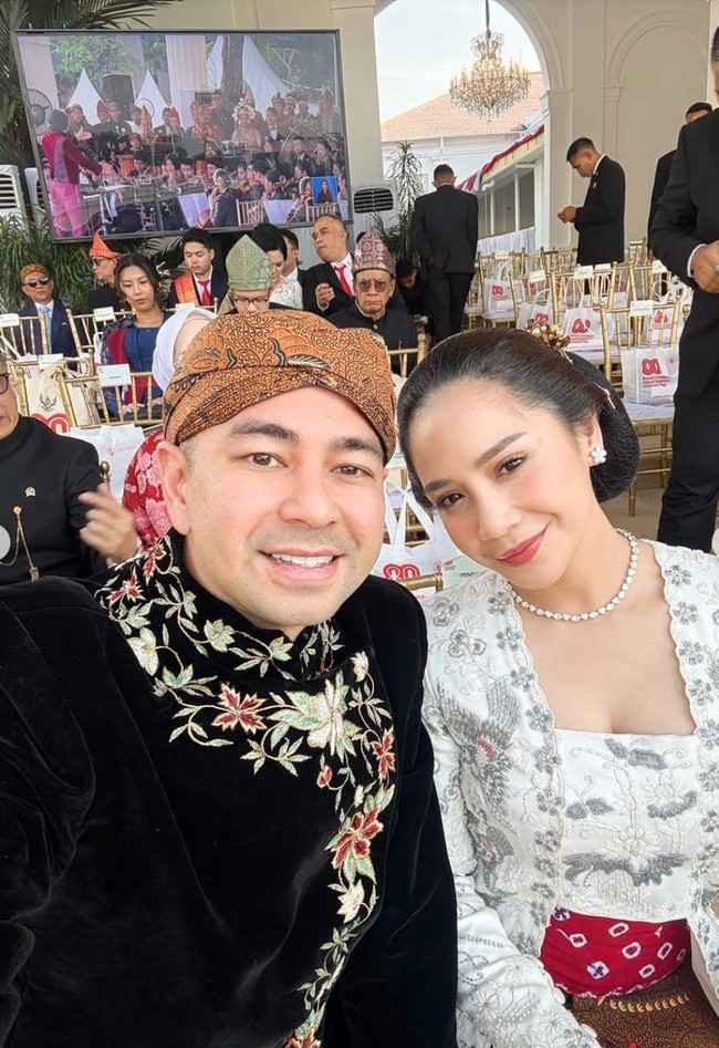 Nagita menyempurnakan penampilannya dengan kalung mutiara sederhana, anting kecil yang menawan, serta handbag putih yang memberi nuansa modern. Rambutnya disanggul rapi dengan hiasan bunga emas kecil di bagian belakang. Foto: Instagram/@raffinagita1717