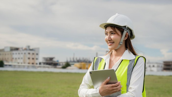 A woman wearing a yellow vest and a hard hat is holding a tablet. She is smiling. The tablet is in her hand