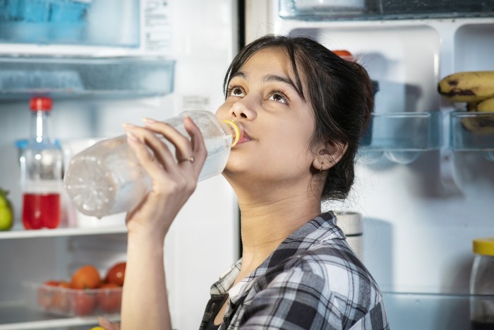Young woman standing in front of open refrigerator and drinking cold water while suffering from a hot summer heat at home