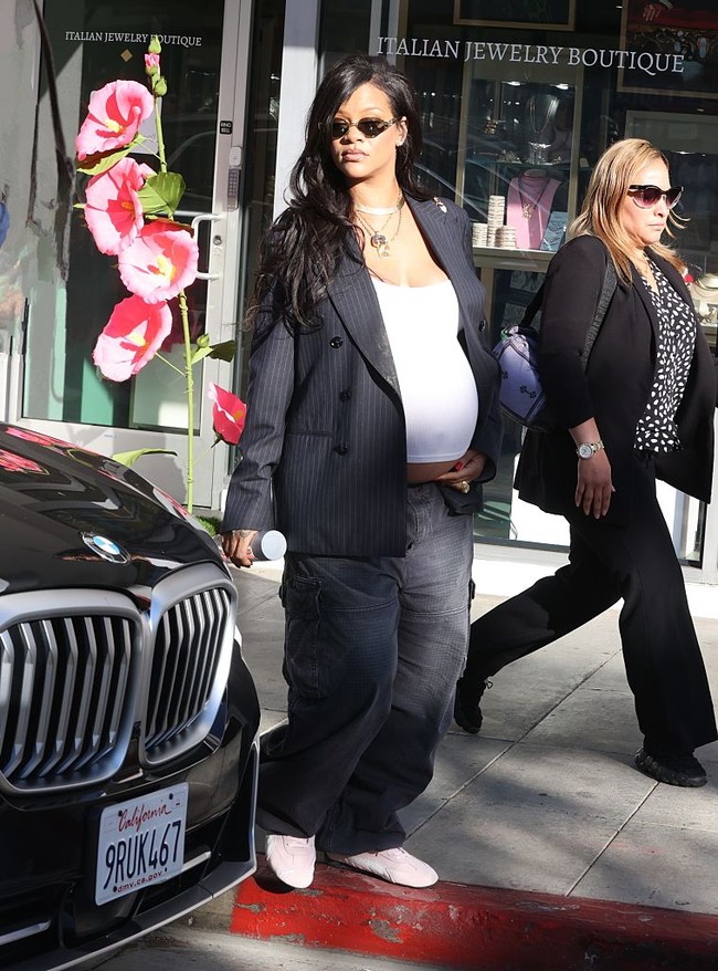 Rihanna mengusung gaya streetwear maternity dengan blazer garis pinstripe oversized dan celana longgar abu-abu. Crop top putih menampilkan perut buncitnya, dipadu sneakers merah muda untuk nuansa santai namun tetap stylish. (Foto: MEGA/GC Images)