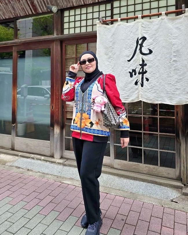 Astrid tampil segar dengan jaket bercorak floral dan motif etnik warna-warni, dipadukan hijab hitam sederhana serta celana hitam. Tas bermotif monogram dengan gantungan boneka pink menambah kesan playful. Foto: Instagram/@astridkuya