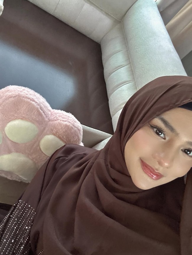 Potret close-up ini menampilkan sisi lembut Davina. Riasan natural dengan sentuhan glossy di bibir, berpadu harmonis dengan hijab cokelat polos yang dililit. Foto: Dok. Instagram/@davinaakaramoy.