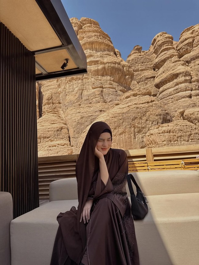 Dalam momen santai, Davina mengenakan hijab pashmina yang ia bentuk syar’i. Duduk di area lounge outdoor, ia memadukan abaya berhias manik-manik halus dengan tas hitam simpel, menciptakan look yang cocok untuk liburan mewah namun tetap tertutup. Hijabnya tetap panjang dan rapi, menyatu dengan warna alam sekitar. Foto: Dok. Instagram/@davinaakaramoy.