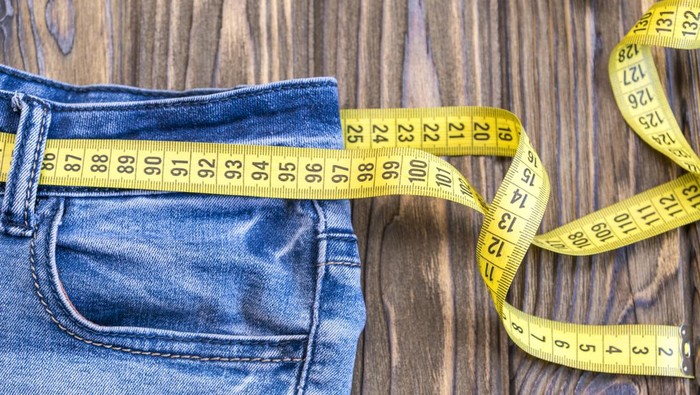 Healthy lifestyle and nutrition concept. Blue jeans with a measuring tape instead of a belt. Close up of jeans with a measuring tape around the waist. Top of the jeans trousers on a wooden background. Diet, sports, weight loss.