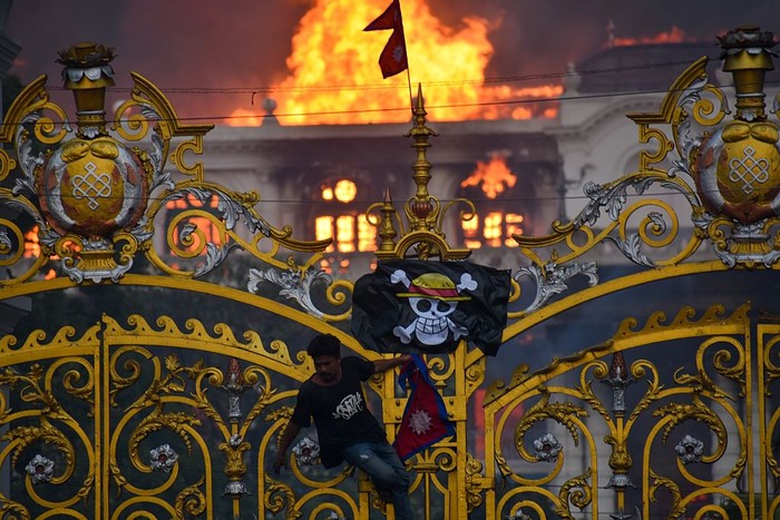 KATHMANDU, NEPAL-SEPTEMBER 9: A man is hanging a pirate flag as smoke and flames rise from the Singha Durbar after people set fire to the Singha Durbar, the seat of Nepal government's various ministers offices in Kathmandu, Nepal on September 9, 2025. At Least 19 people were killed and dozens injured on September 8 during the demonstration against corruption and ban on social media by the government. (Photo by Sunil Pradhan/Anadolu via Getty Images)