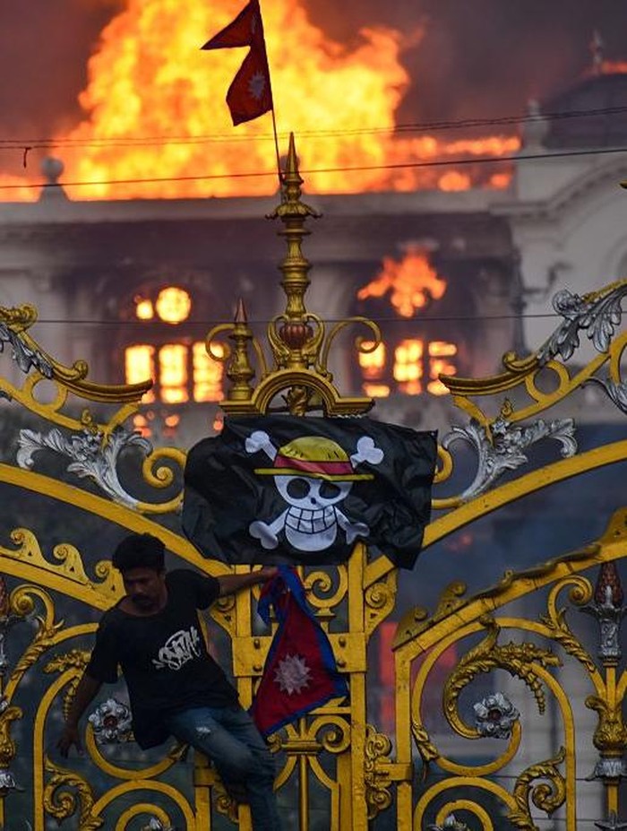 Bendera Jolly Roger One Piece Saat Demonstrasi di Nepal KATHMANDU, NEPAL-SEPTEMBER 9: A man is hanging a pirate flag as smoke and flames rise from the Singha Durbar after people set fire to the Singha Durbar, the seat of Nepal governments various ministers offices in Kathmandu, Nepal on September 9, 2025. At Least 19 people were killed and dozens injured on September 8 during the demonstration against corruption and ban on social media by the government. (Photo by Sunil Pradhan/Anadolu via Getty Images)