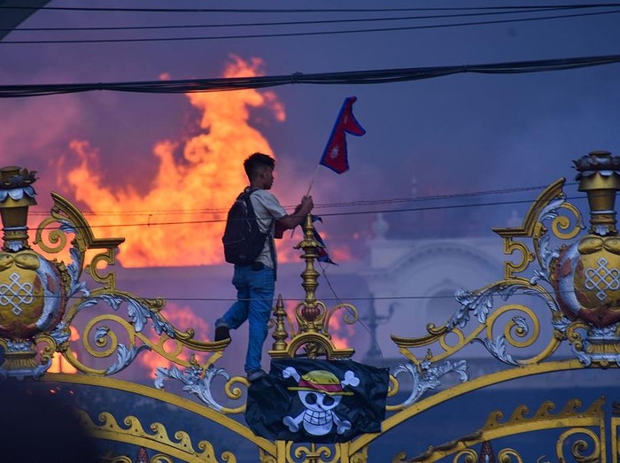 Bendera Jolly Roger One Piece Saat Demonstrasi di Nepal KATHMANDU, NEPAL-SEPTEMBER 9: A man is hanging a pirate flag as smoke and flames rise from the Singha Durbar after people set fire to the Singha Durbar, the seat of Nepal governments various ministers offices in Kathmandu, Nepal on September 9, 2025. At Least 19 people were killed and dozens injured on September 8 during the demonstration against corruption and ban on social media by the government. (Photo by Sunil Pradhan/Anadolu via Getty Images)