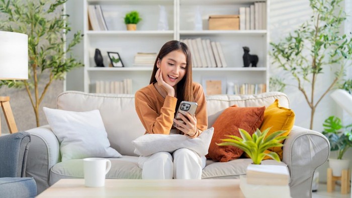 Smiling Asian cheerful adult female enjoy using smartphone surf internet social media relax casual daily technology in life,woman use smartphone weekend on wooden bench near window in living room at home