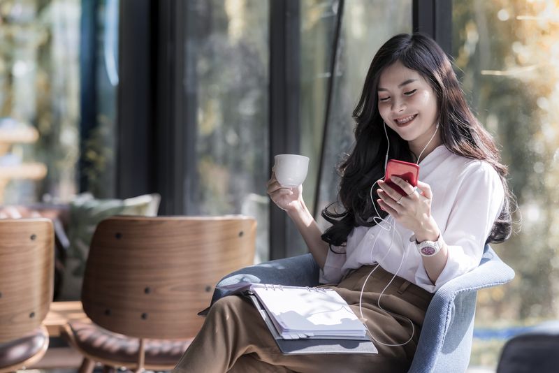 Shot of a beautiful young asian woman holding cup of coffee and listening  music from her phone in the coffee shop