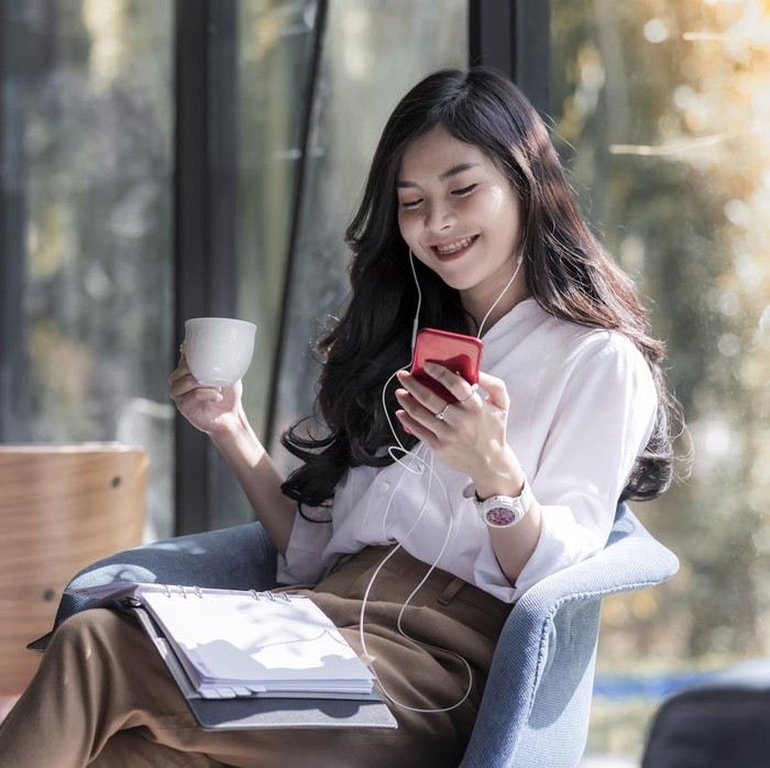 Shot of a beautiful young asian woman holding cup of coffee and listening  music from her phone in the coffee shop