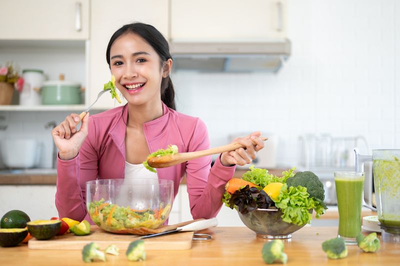 An attractive and cheerful Asian woman in sportswear enjoys her healthy salad at a kitchen table in the kitchen. Healthy lifestyle concept
