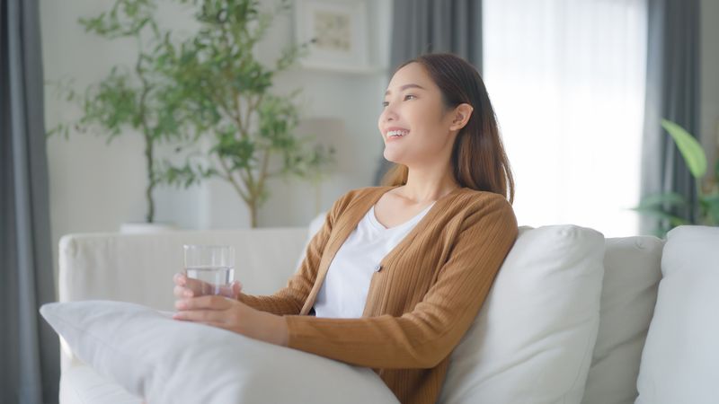 Happy young Asian woman drinking water while sitting on the sofa in the living room