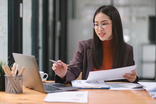 Asian businesswoman or accountant using a calculator, graphs, and charts to analyze market data, balance sheets, accounts, and net profits