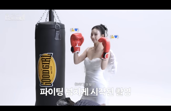 Karina aespa tampil memukau dengan gaun pengantin off-shoulder berwarna putih, tatanan rambut model bun, serta sayap di belakang punggungnya. Namun, ia juga memakai sarung tangan tinju berwarna merah dan syuting adegan melakukan pukulan ke samsak. Foto: dok. SM Entertainment