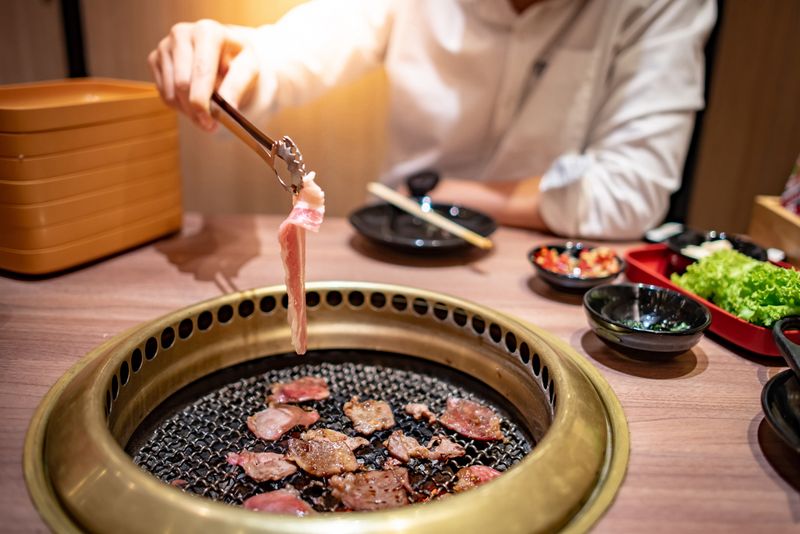 Ilustrasi restoran atau tempat makan Asian man using kitchen tongs grilling beef and bacon on grill plate. Eating Korean Barbecue buffet in the restaurant.