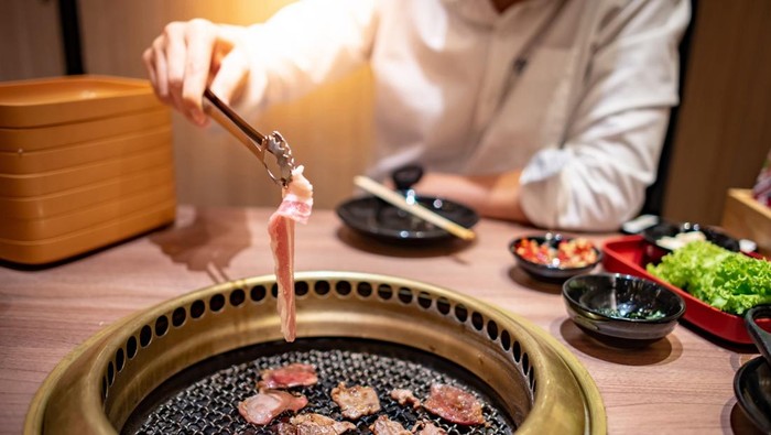 Ilustrasi restoran atau tempat makan Asian man using kitchen tongs grilling beef and bacon on grill plate. Eating Korean Barbecue buffet in the restaurant.