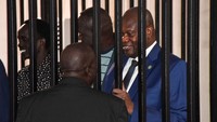 Wakil Presiden Pertama Sudan Selatan yang diskors, Riek Machar, berdiri di balik jeruji baja ruang persidangan, saat ia berbicara dengan Dr. Geri Raimondu Lege, dari tim pembelanya, selama persidangan atas tuduhan termasuk pengkhianatan dan kejahatan terhadap kemanusiaan, di Freedom Hall di Juba, Sudan Selatan, Rabu (24/9/2025). REUTERS/Jok Solomun