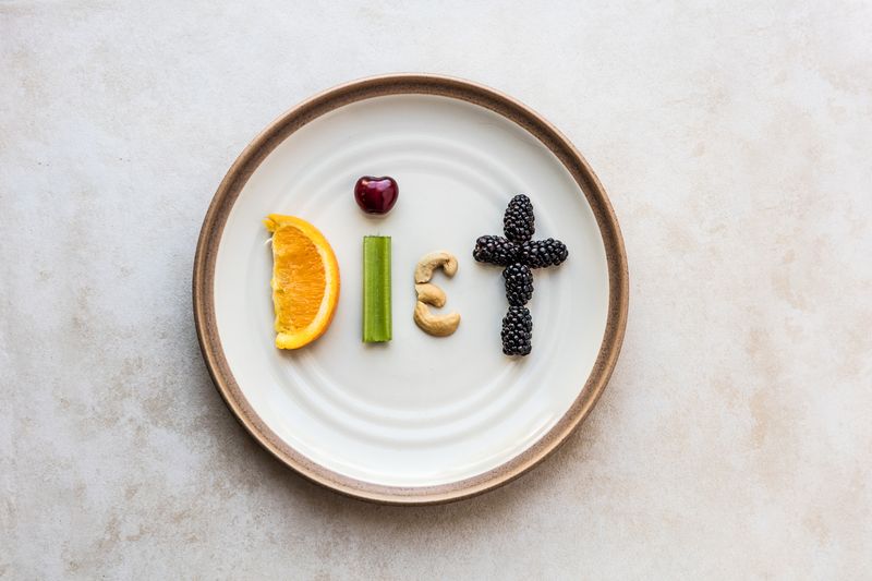 The word Diet made from food. A orange slice, celery stick, cherry, cashew, and blackberry spell diet, as it sits on a stoneware plate. Using a flat lay over head view to capture the spelled out food word.