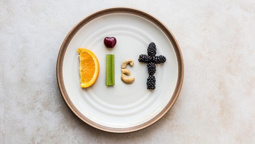 The word Diet made from food. A orange slice, celery stick, cherry, cashew, and blackberry spell diet, as it sits on a stoneware plate. Using a flat lay over head view to capture the spelled out food word.