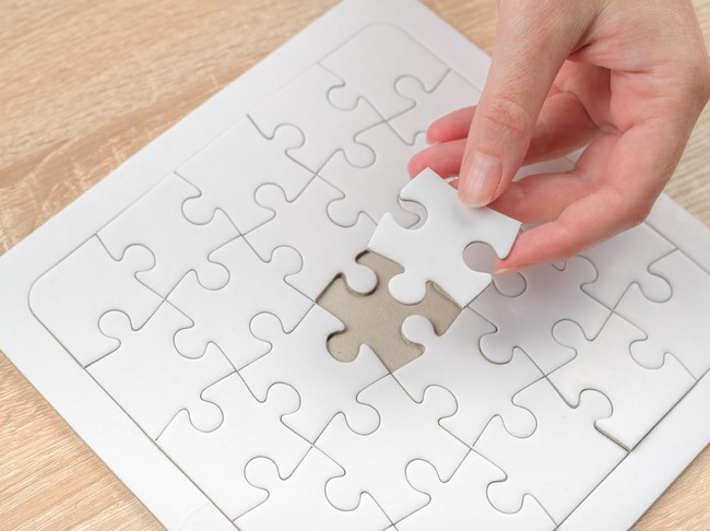 Female hand putting a missing piece and solving blank white jigsaw puzzle placed on top of old wooden oak table, close up, selective focus