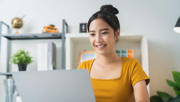 Happy young Asian woman using laptop and checking email inbox, social media messages on the table in modern home.