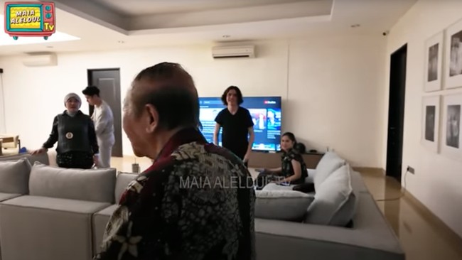 Ruang keluarga dilengkapi dengan sofa besar berwarna abu-abu yang nyaman, dengan TV layar lebar menempel di dinding. Ruangan tersebut terasa hangat dengan pencahayaan yang pas dan dominasi warna coklat. Foto: YouTube/MAIA ALELDUL TV