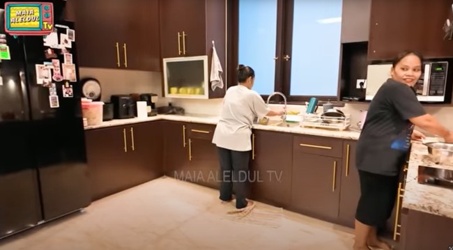 Dapur terlihat modern dengan kabinet berwarna cokelat tua dan pegangan emas yang elegan. Peralatan dapur seperti microwave, kulkas, dan mesin kopi juga lengkap tersedia. Foto: YouTube/MAIA ALELDUL TV