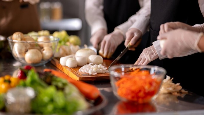 Close up of gloved hands chef holding knife chopping onions on cutting board at kitchen counter full of vegetables. Cooking Class, Food preparation.
