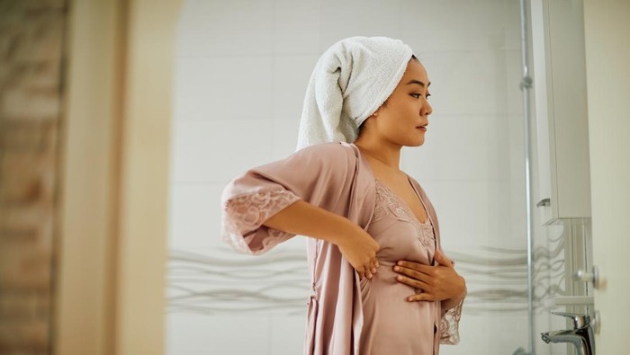 Young Asian woman standing in front of bathroom mirror while doing breast self-examination. Copy space.