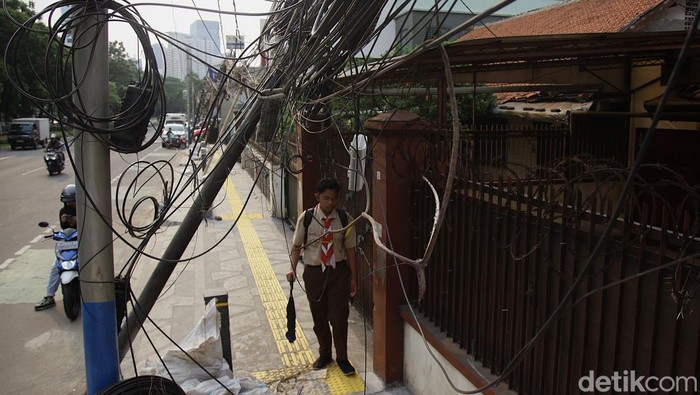 Warga melintas di antara kabel fiber yang menjuntai dari tiang yang miring dan nyaris ambruk di Jl Abdullah Syafei, Tebet, Jakarta Selatan, Rabu (8/10/2025). Tampak bagian bawah berupa jalur pedestrian yang masih dalam tahap akhir revitalisasi (belum selesai sepenuhnya).