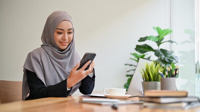Beautiful muslim businesswoman or female executive manager wearing hijab sits at her worktable and uses a smartphone.
