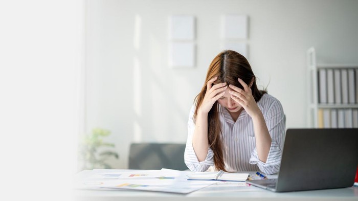A woman is sitting at a desk with a laptop and a stack of papers. She is looking at the laptop and she is in a state of distress