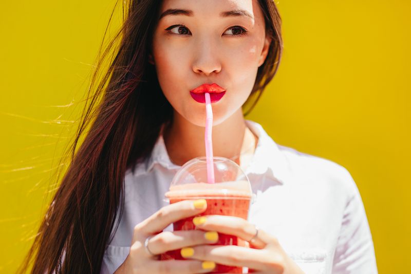 Asian woman drinking juice in a disposable plastic glass using a straw. Portrait of a young woman enjoying a glass of juice.