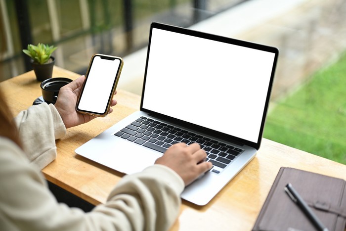 Unrecognizable woman holding mobile phone while using laptop on wooden table.