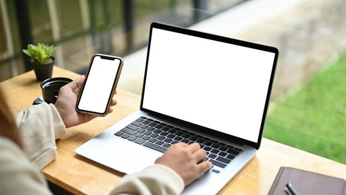Unrecognizable woman holding mobile phone while using laptop on wooden table.