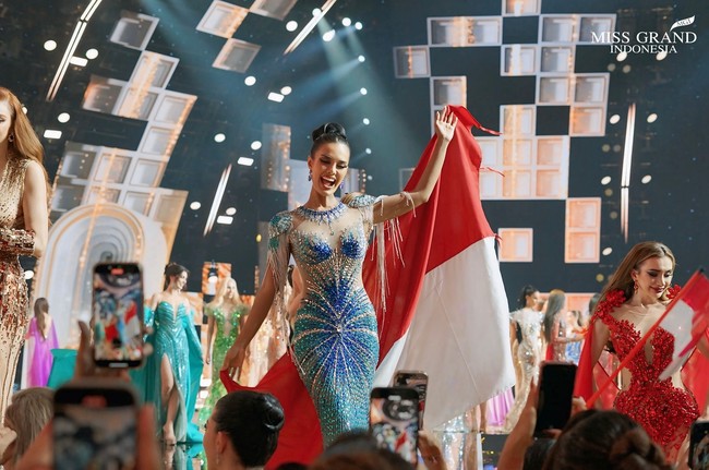 Lolos hingga Top 22, Vina menutup perjalanannya di Miss Grand International 2025 dengan gaun berwarna biru dan nude berhiaskan ribuan kristal rancangan Herry Nadjaya. Memegang bendera Merah Putih di tangan, penampilannya menjadi simbol kebanggaan dan dedikasi terhadap Tanah Air. Foto: Instagram/@vinanggisitorus